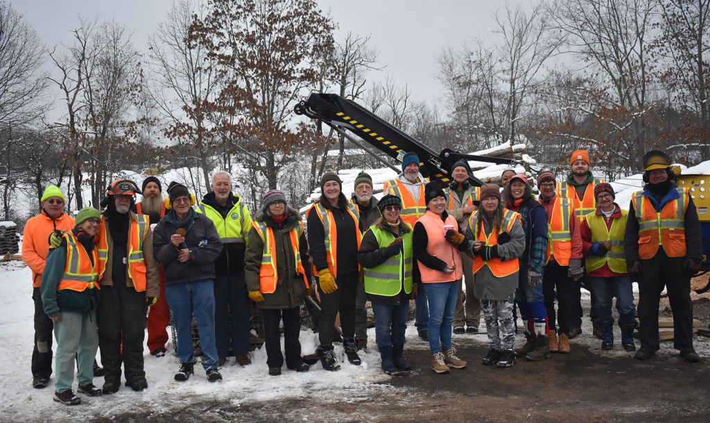 Group of about 20 people geared up with safety vests in front of firewood equipment.