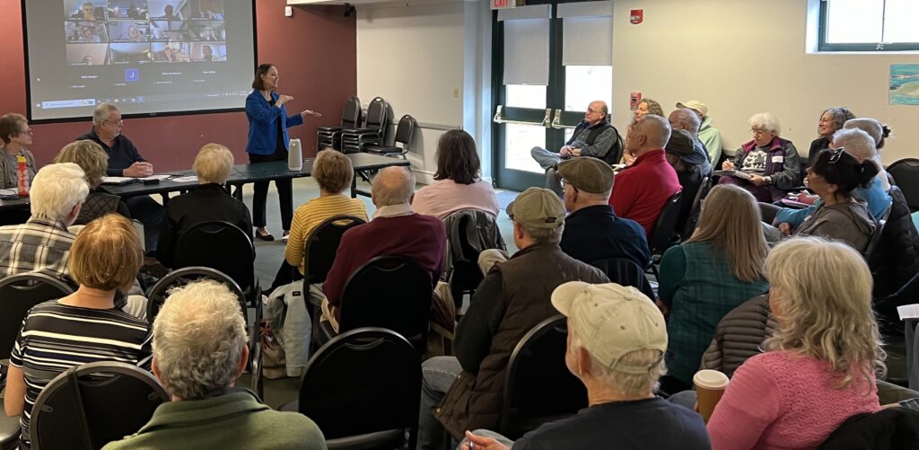 Secretary of State Shenna Bellows speaks to packed room of Waldo County Dems with Zoom attendees in background